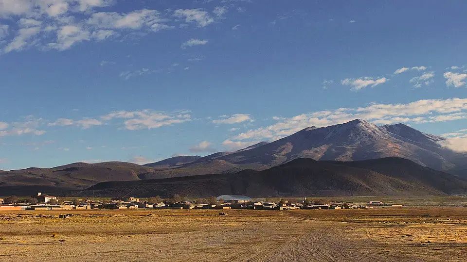 A rural community in the Bolivian altiplano: low adobe and brick houses spread across a dry plain, with dark mountains and a snow-dusted peak beyond